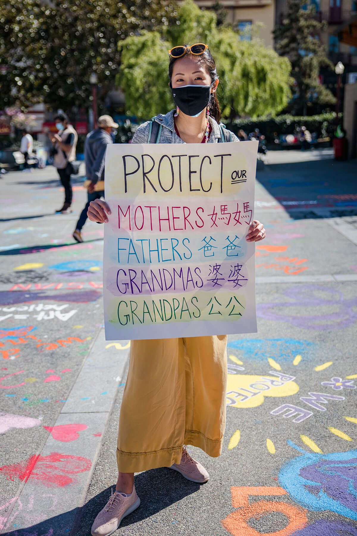 Asian woman standing at a Stop Asian Hate protest holding a sign that says, "Protect our Mothers, Fathers, Grandmas, and Grandpas." Each word also has its Chinese translation.