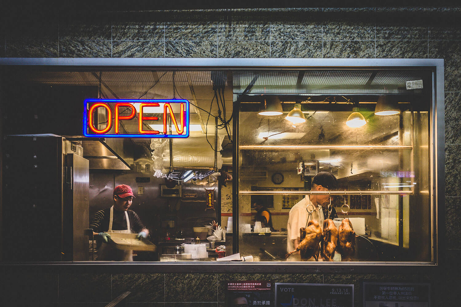 Two people standing in the kitchen of a restaurant in Chinatown, New York. Two roasted ducks and a glowing neon Open sign hang in the window.