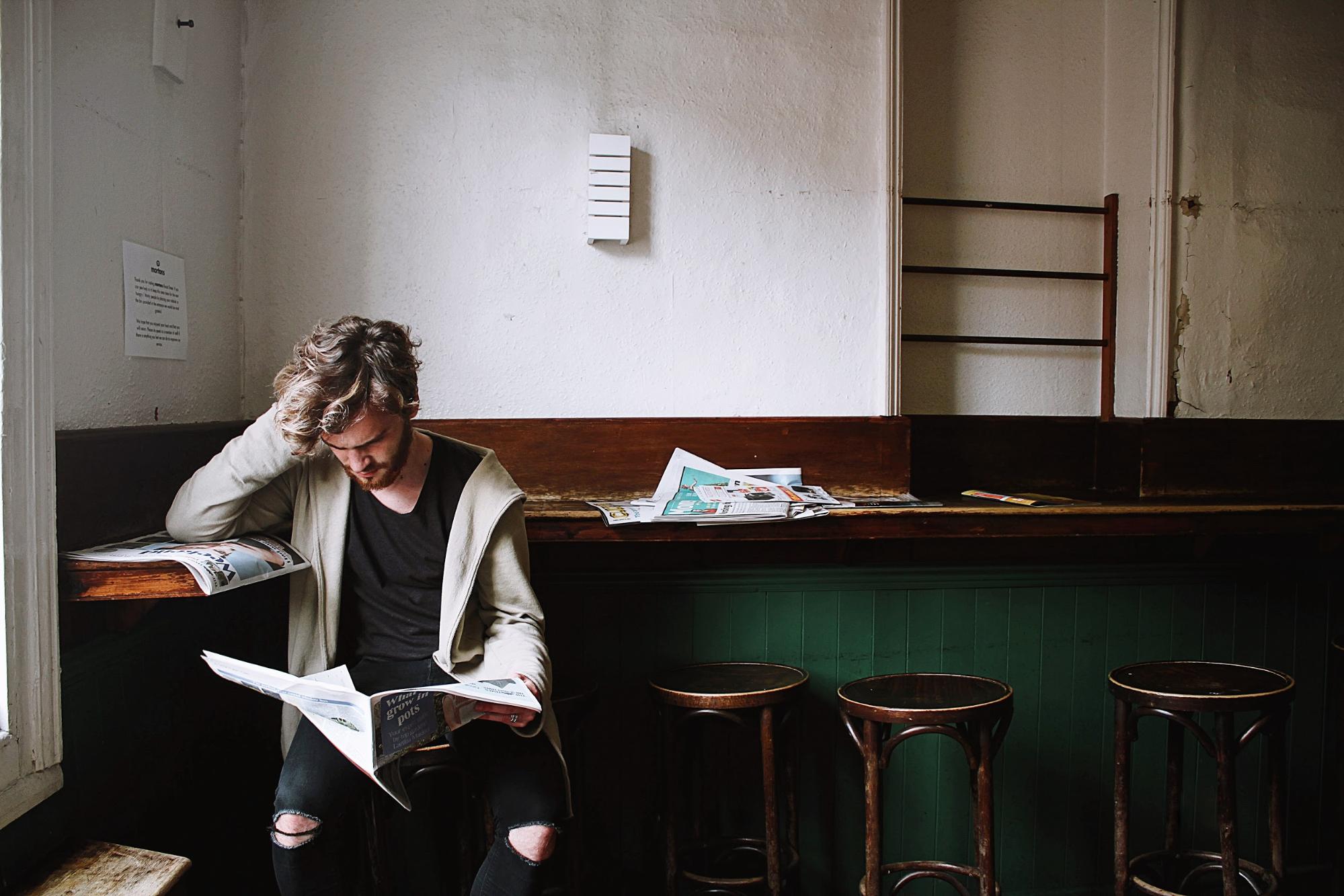 A man sitting on a stool in a coffee shop reading the newspaper. He has a look of concentration.