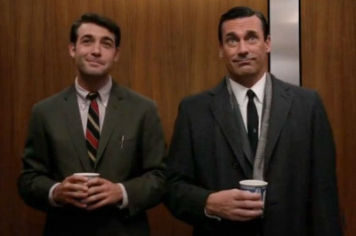 Don draper on an elevator with a colleague. Both in suits holding coffee and smiling.
