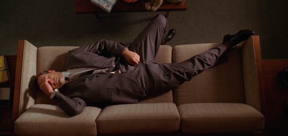An aerial view of Don Draper laying on a couch in a suit. His hand is pressed to his forehead and he looks tired and stressed.