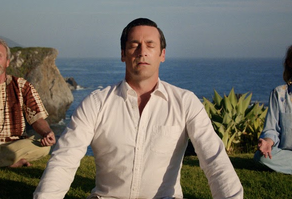 Don Draper meditating on a grassy lawn with the ocean in the background.