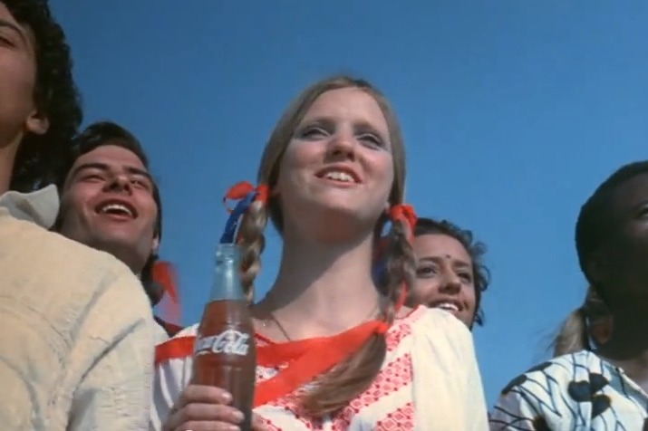 A close up of a woman in a crowd looking happy and holding a bottle of Coca Cola. Circa 1970s.
