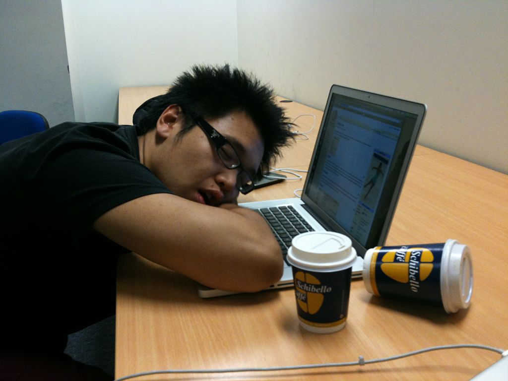 A person asleep at a desk with their head on their laptop. Two coffee cups sit on the desk beside them, one toppled over.