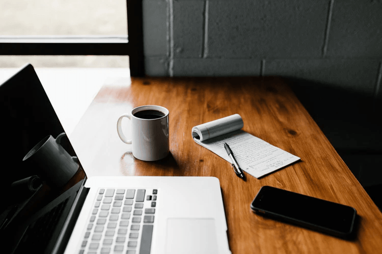 A computer, coffee mug, phone, and pad of paper sitting on a wooden table.