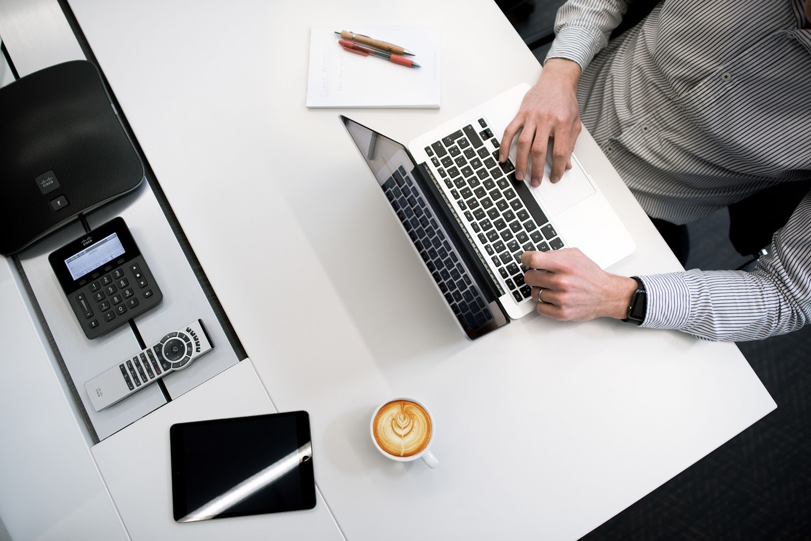 man working on desk with phone, remote, tablet, laptop, and coffee