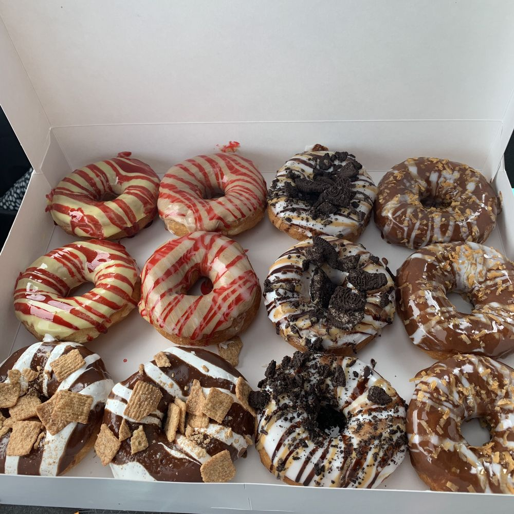 A box of donuts from Dough-Hio Donuts in Lancaster, OH, including yellow donuts with yellow icing and red drizzle, white icing with oreo crumbs, and chocolate icing with cinnamon toast crunch cereal.