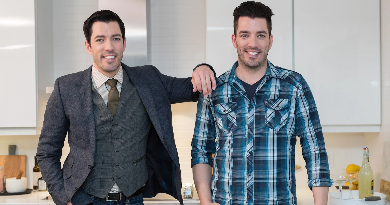 Drew Scott in a three-piece suit and Jonathan Scott in a flannel button-down stand side by side in immaculate white kitchen