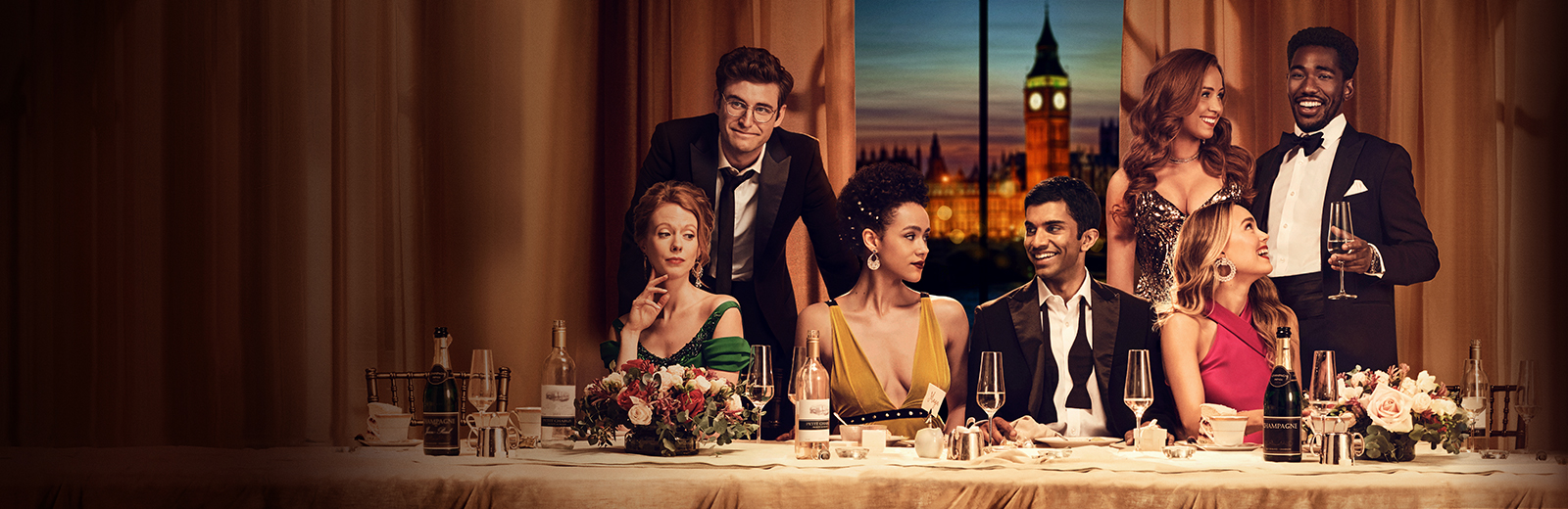 The cast of Four Weddings and a Funeral sit and stand around a fancy dining table set with champagne and flowers