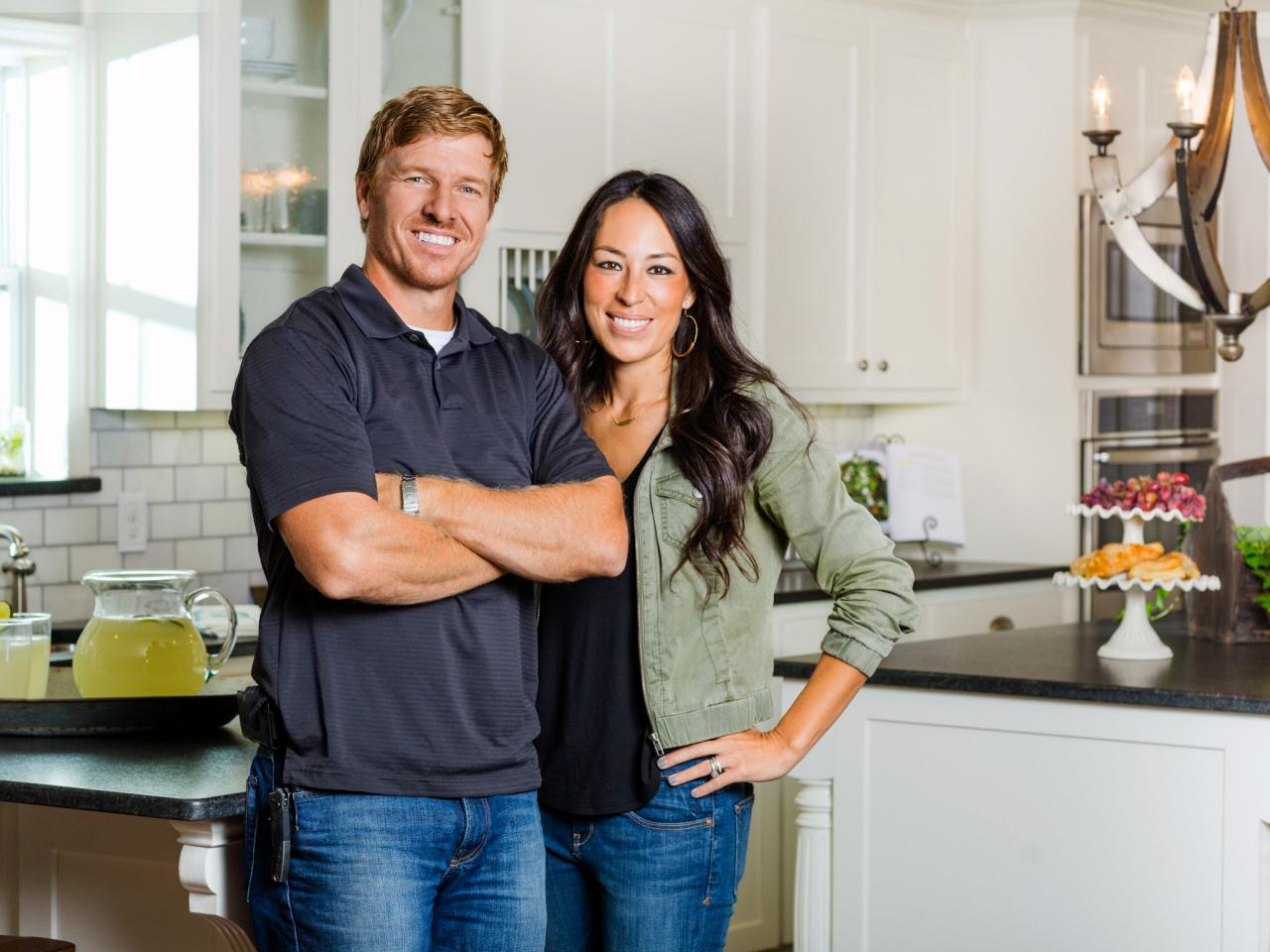 Joanna and Chip Gaines stand side by side smiling in an immaculate white and black kitchen