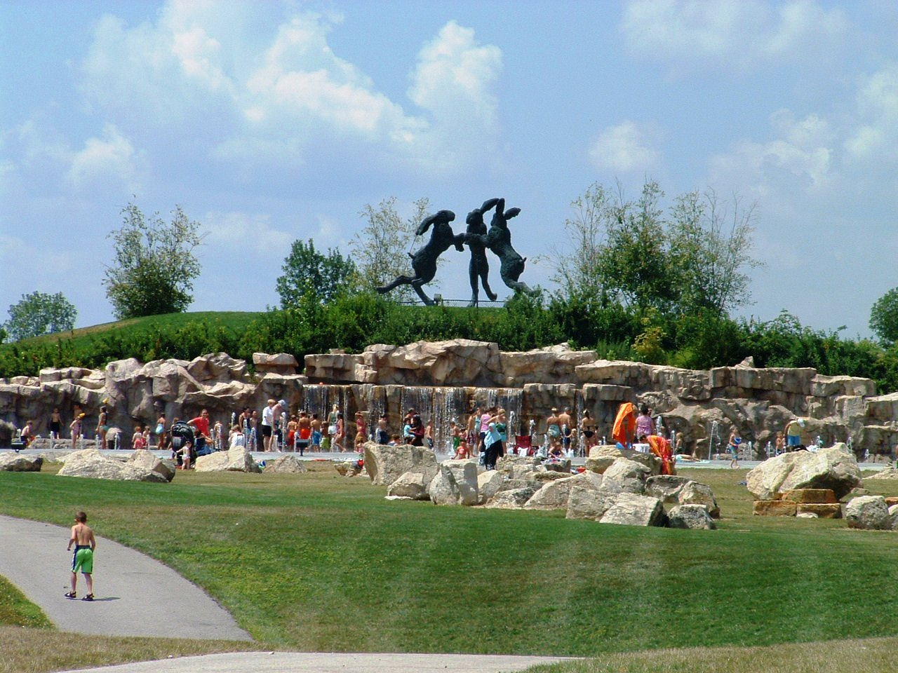 kids playing in a large water fountain in front of large statue of three dancing hairs