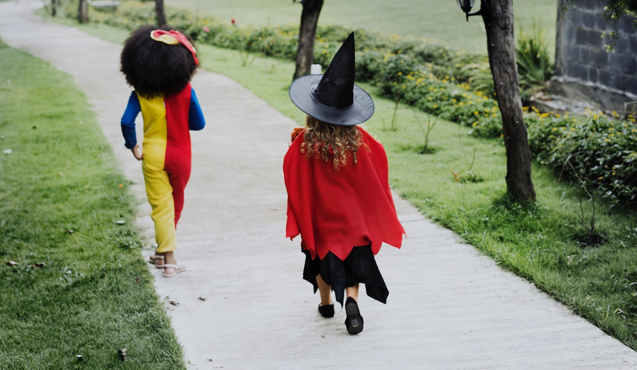 A child in a clown costume and a child in a which costume run down a sidewalk, excited to trick or treat on halloween.