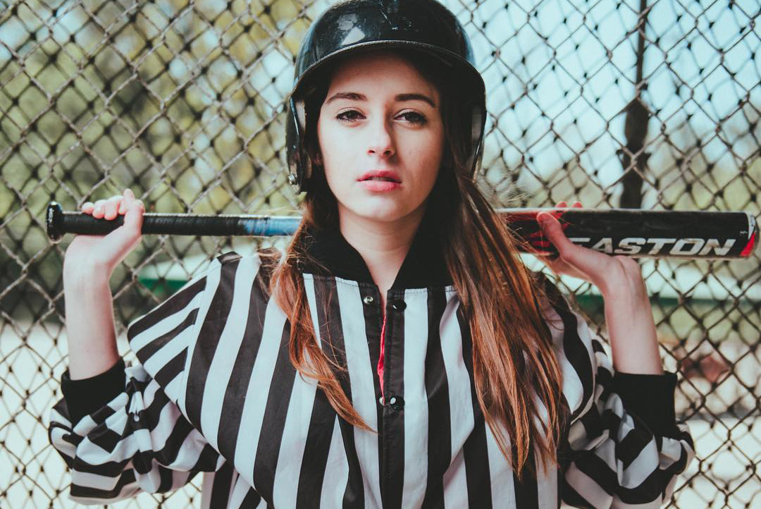 Young woman in batting cage with helmet on and bat around the bak of her neck. Redhead.