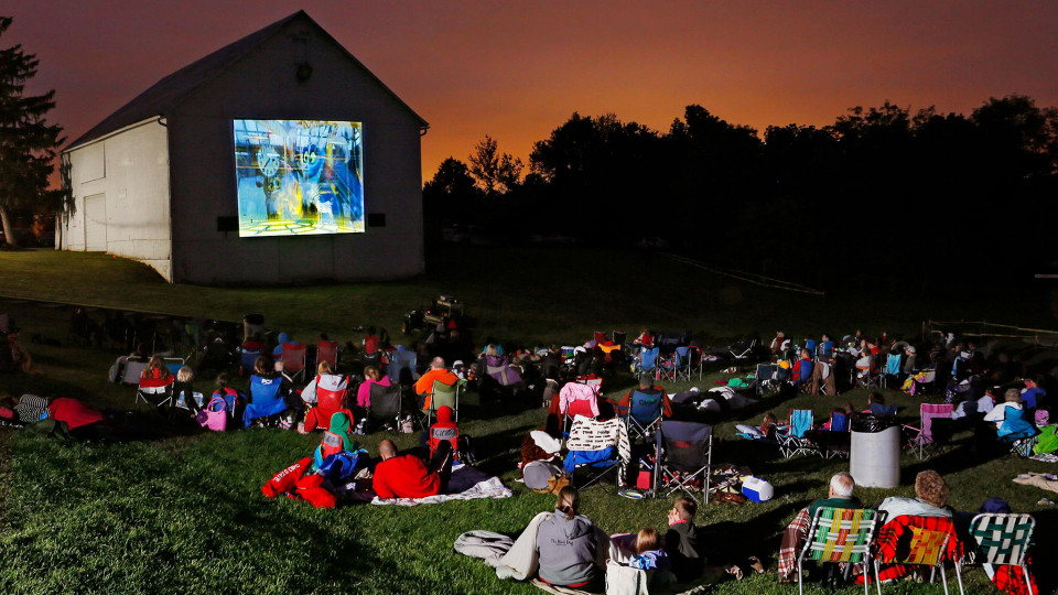 A movie is being projected on the side of a building as a large crowd rests in a lawn watching. 