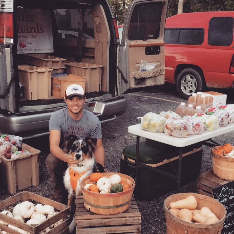 A man and his dog sit behind a table of bagged apples at a farmers market