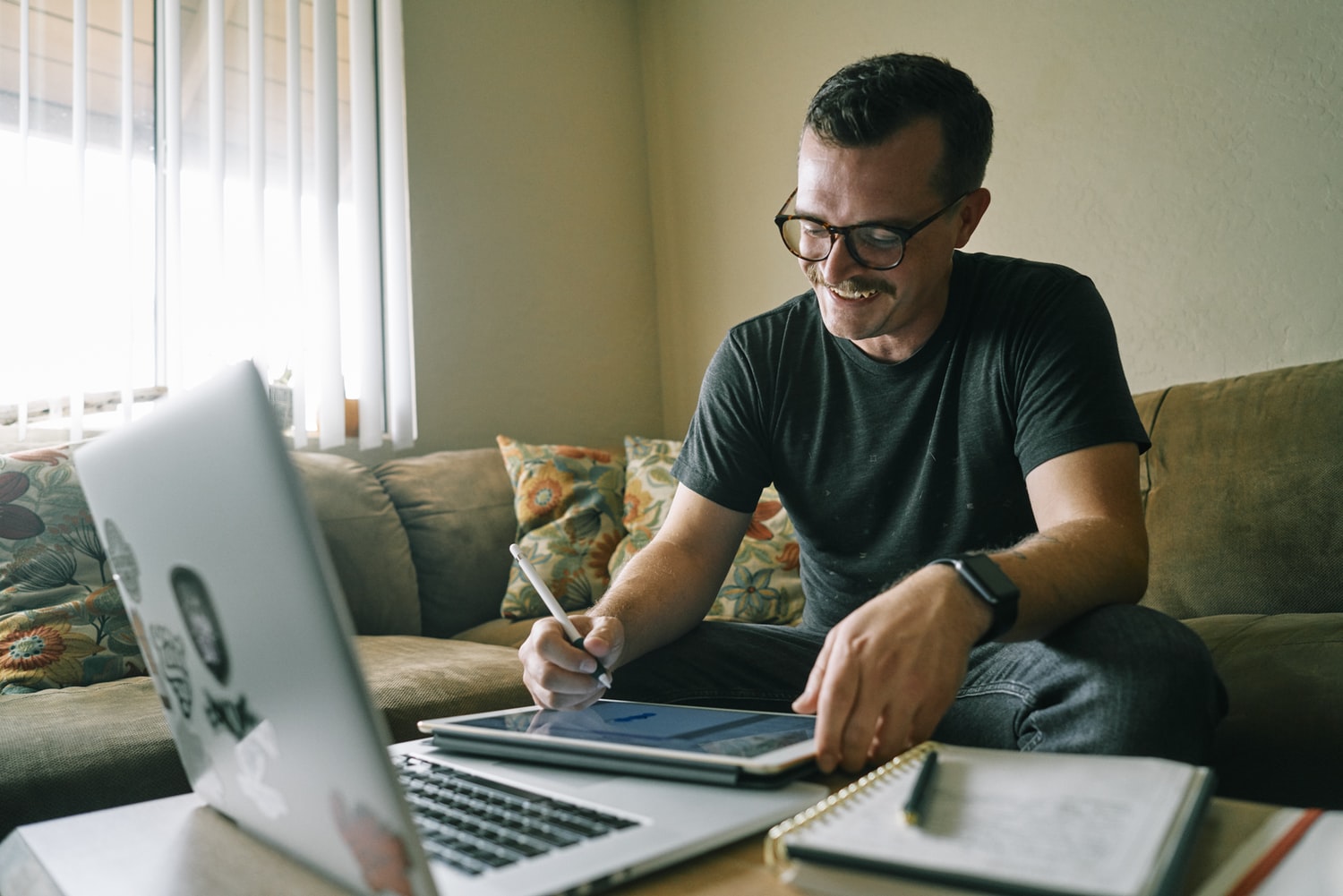 A man smiling and writing.  He is working from home on his couch, with a laptop in the foreground.