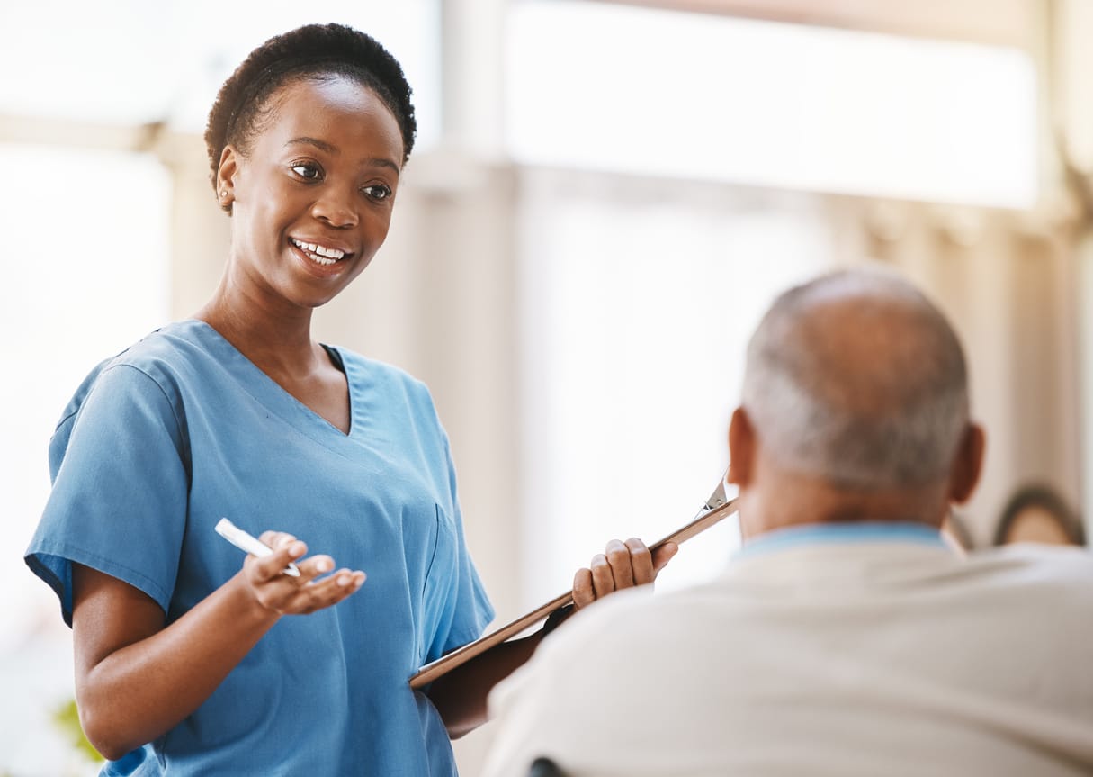 A mental health tech in scrubs holds a clipboard and speaks with a patient.