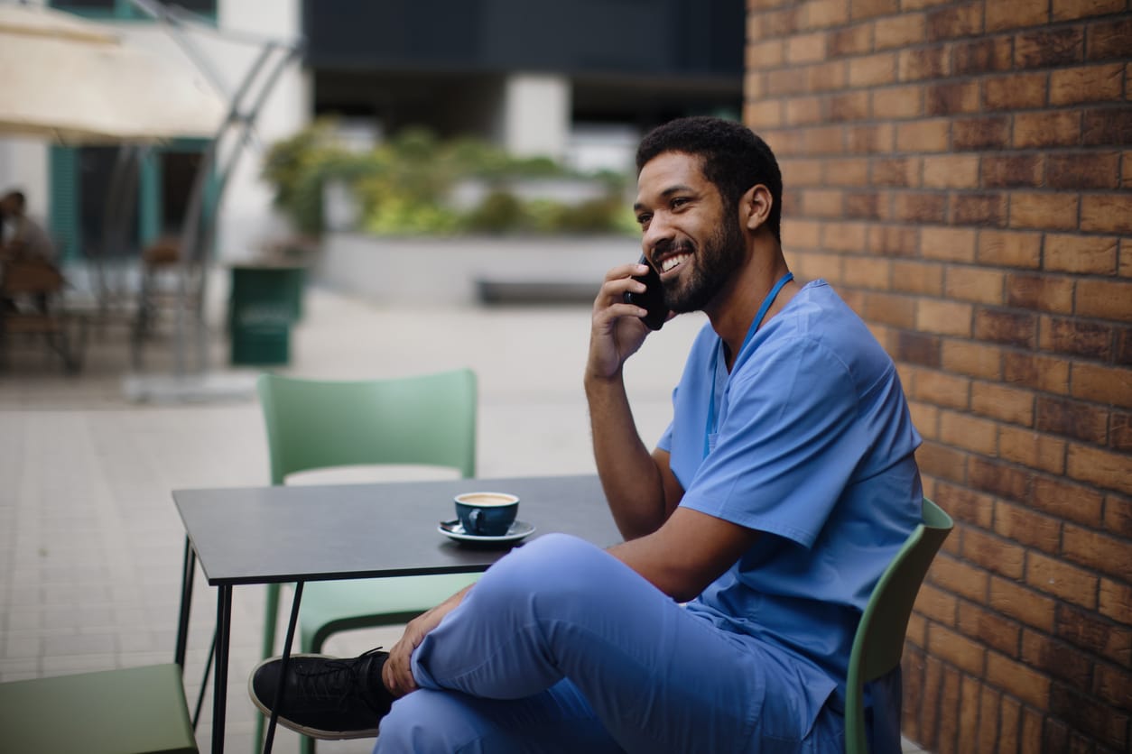 A nurse in blue scrubs sits outside at a cafe with a coffee talking on his phone.