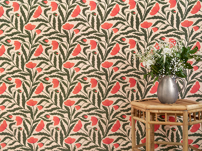 Wallpaper with beige background and a dark green and coral poppy pattern. Rattan side table in the foreground with a silver pitcher and white and green flowers inside.