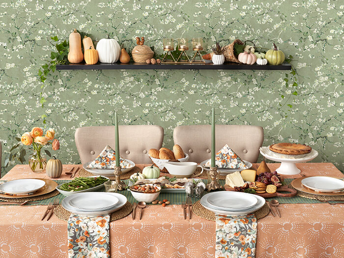 Thanksgiving table set with a clay-orange tablecloth, floral napkins, and green tapered candles. The table is set with plates, silverware, and a plate of cheese and crackers and green beans is in the center waiting to be enjoyed. There is green floral wallpaper in the background and the vibes are festive and Autumnal complete with white, orange and green pumpkins and other fall decor.