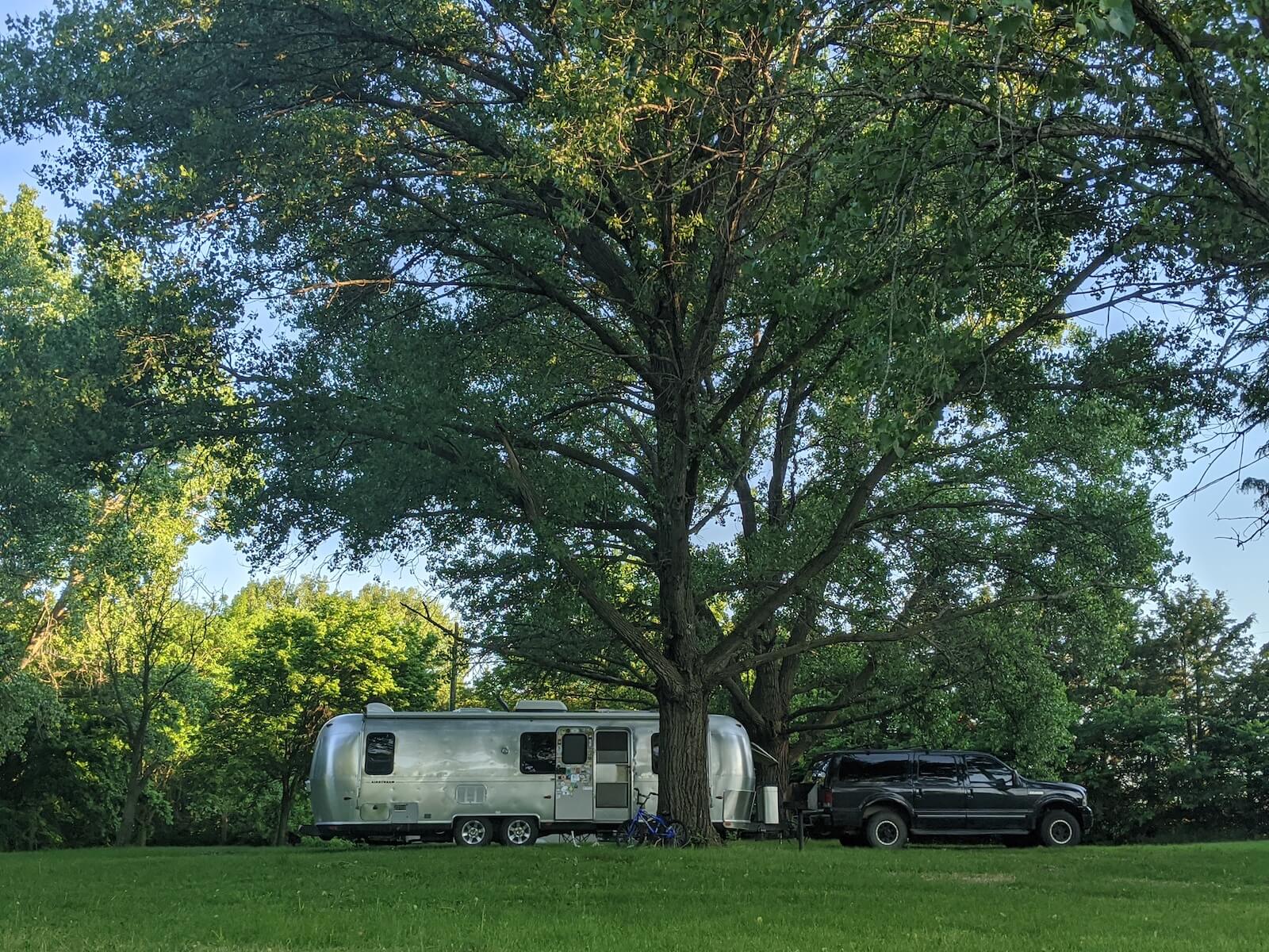 large truck and airstream rv parking underneath large tree with grassy field in the foreground travel nursing in an RV