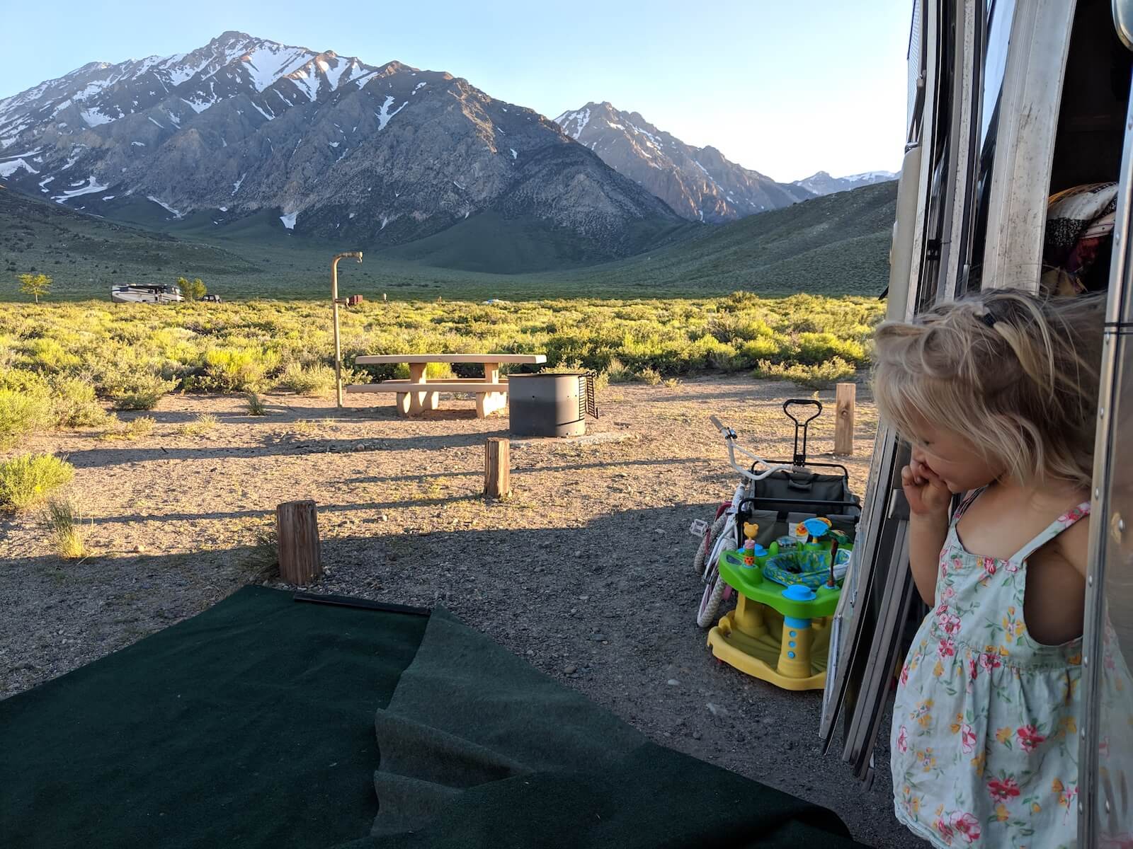 little girl stepping out of airstream door with mountains and grassy field in the background travel nursing in an RV