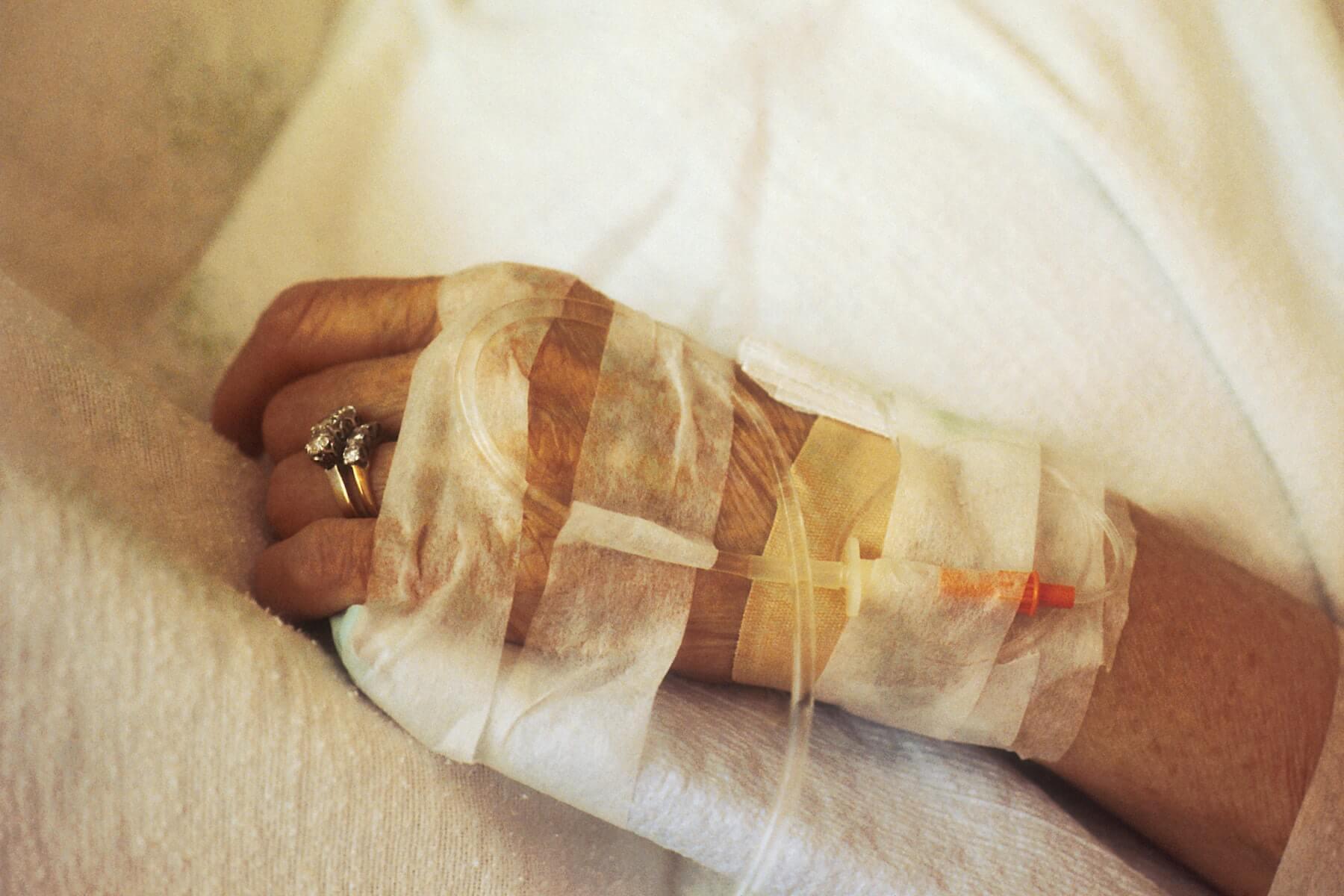 hand of old woman with IV drop attached and wedding ring on finger laying on side of bed hospice nursing