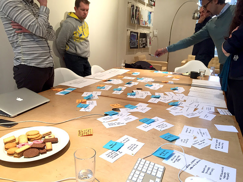 A card sorting workshop in progress. People stand around a table that's full of cards and post-it notes. Photograph.