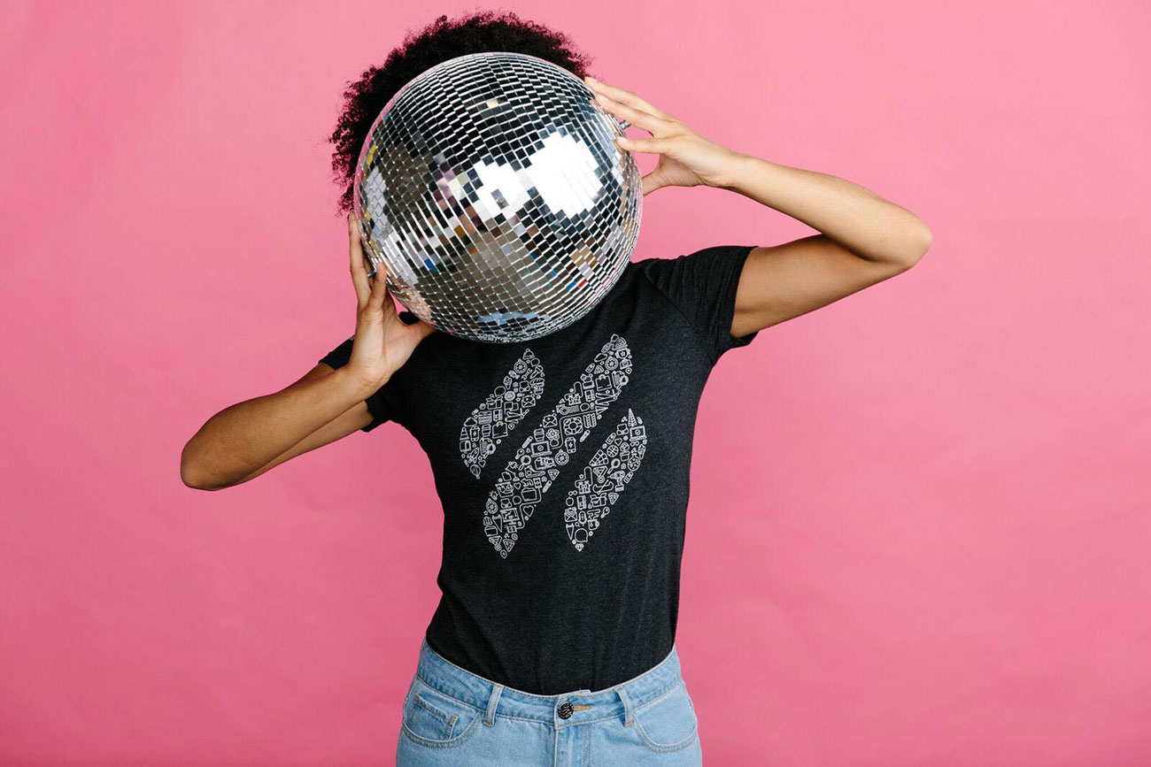 Woman holding disco ball in front of face wearing a black shirt and jeans in front of a pink background