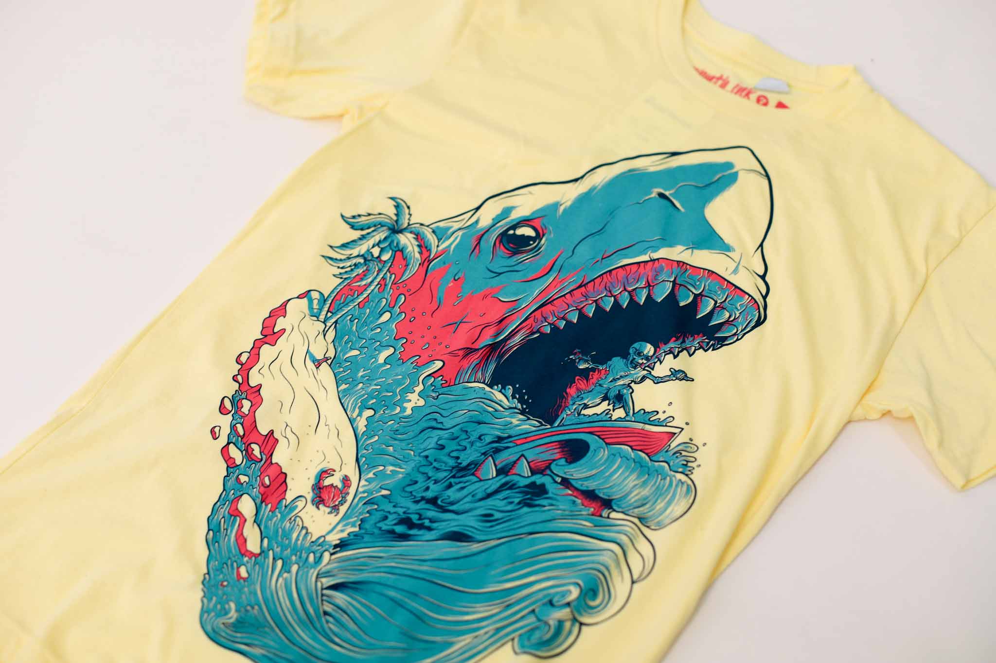 Yellow shirt with artwork of blue and pink shark with skeleton surfing in its mouth