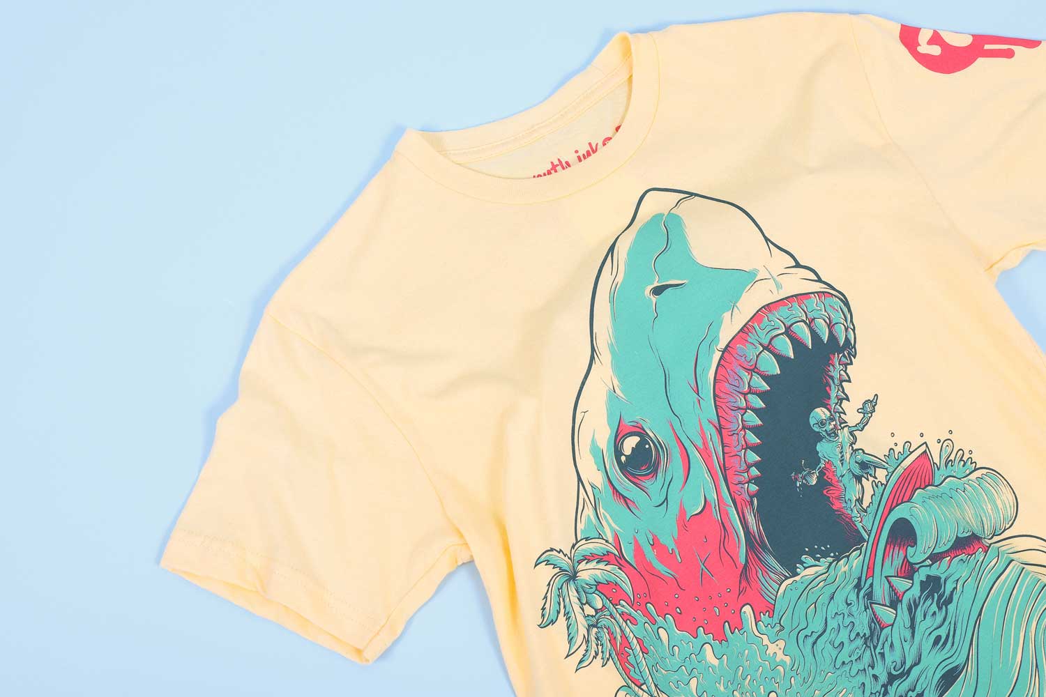 yellow shirt with blue and pink stylized shark and a skeleton riding a surfboard out of its mouth