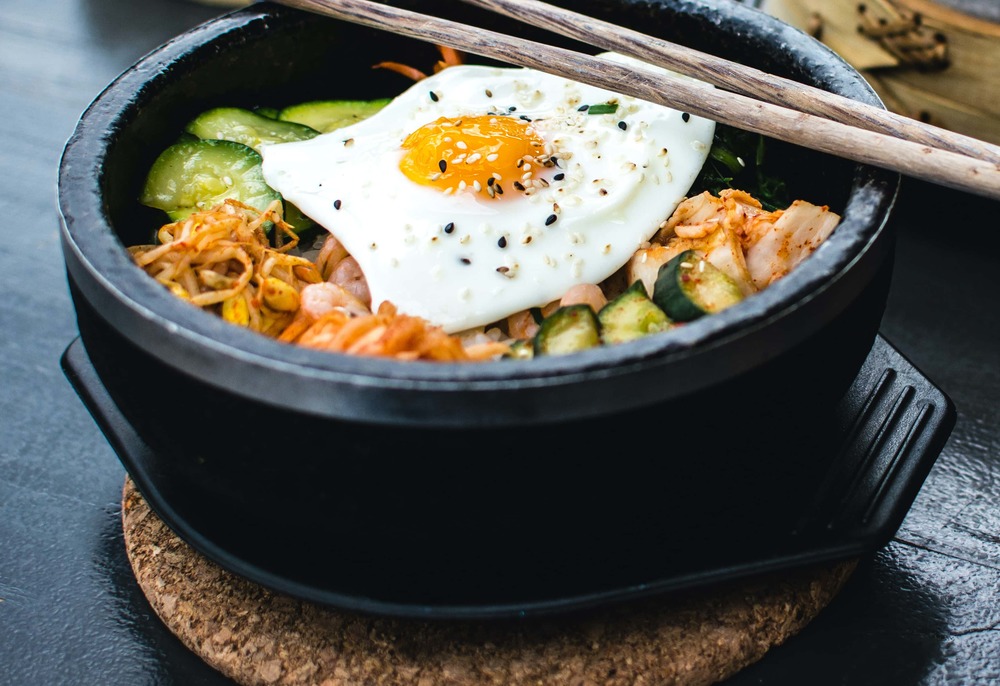 Dans un grand saladier en fonte, on retrouve l'ensemble des ingrédients qui composent ce bibimbap. Un oeuf au plat est sur le dessus.