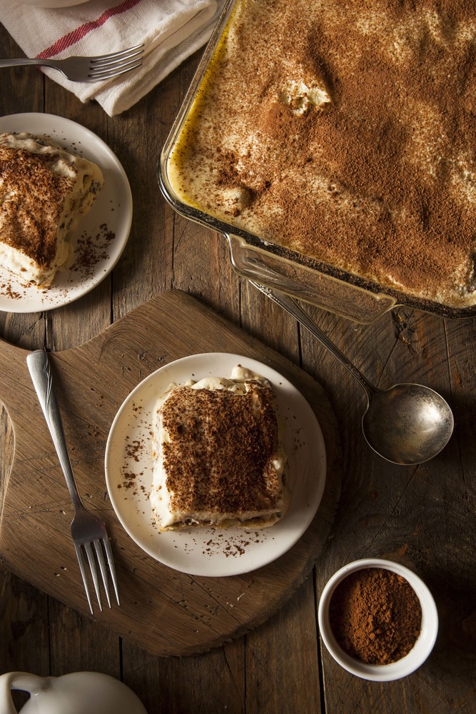 Le tiramisu protéiné est disposé dans un grand moule transparent. Deux parts sont déjà dans des assiettes à dessert, prêtes à être consommés. 