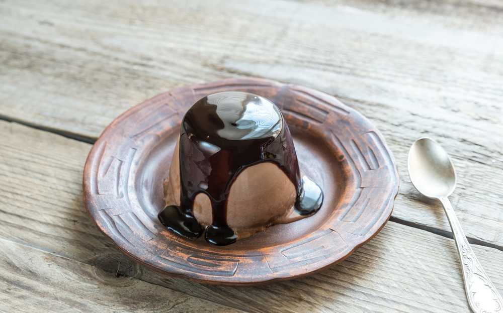 Posée sur une table en bois clair, une assiette creuse de couleur beige accueille la Panna cotta recouverte par un chocolat coulant. 