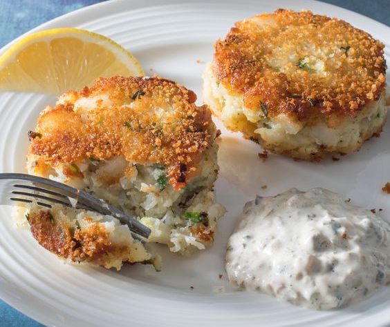 Deux grosses croquettes de cabillaud sont déposées sue une assiette de couleur blanche. Une tranche de citron jaune est présente.