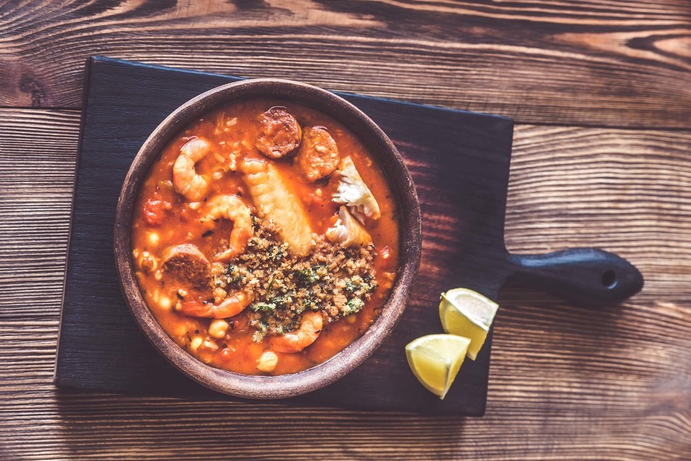 Sur une planche à découper, un bol de couleur marron est posé avec à l'intérieur la soupe de la mer. Deux quartiers de citron jaune sont à côté.