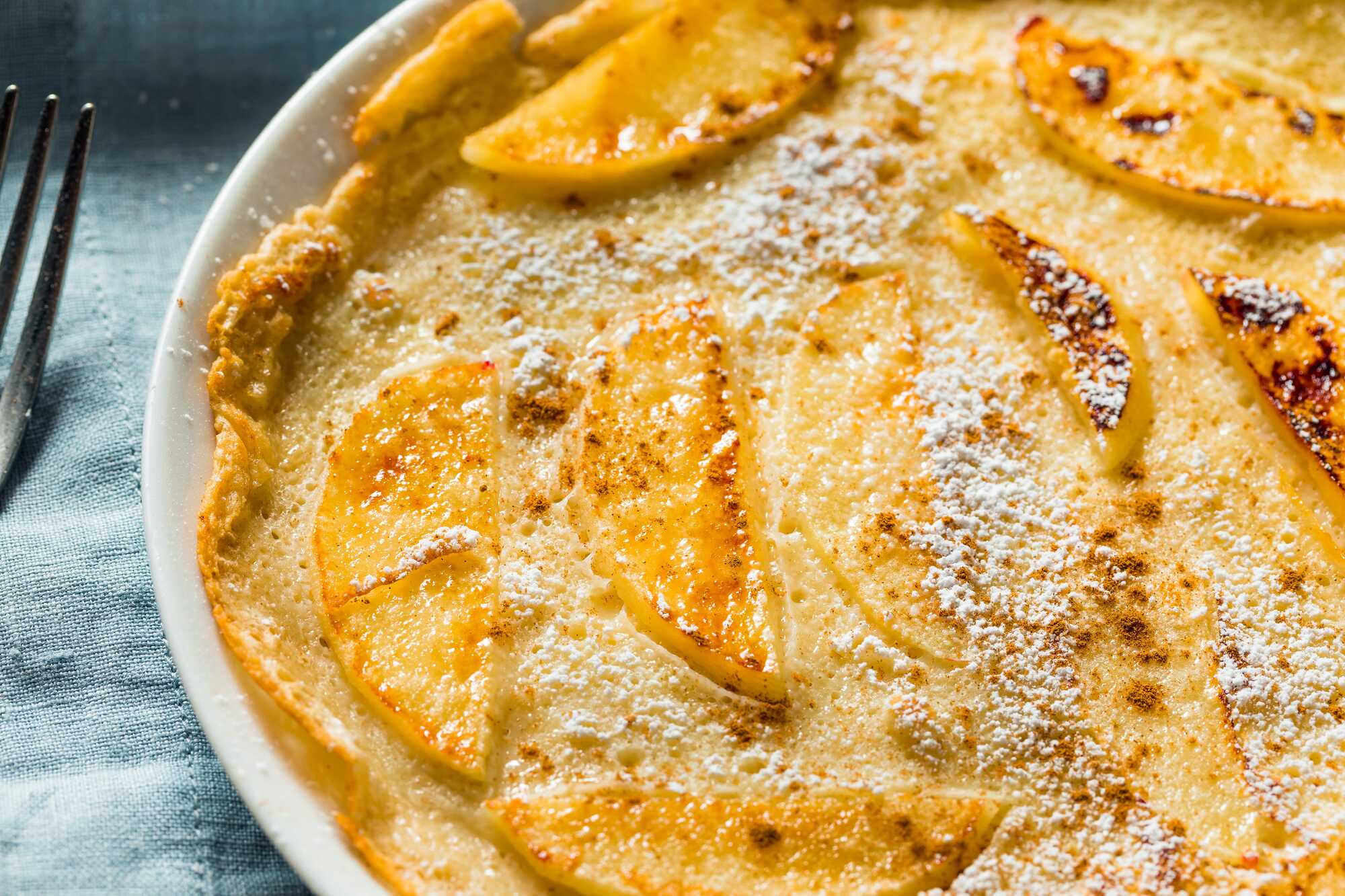 Dans un plat à tarte blanc, se trouve la maxi crêpe protéinée. Les pommes soigneusement réparties dans le plat sont coupées en tranches généreuses.