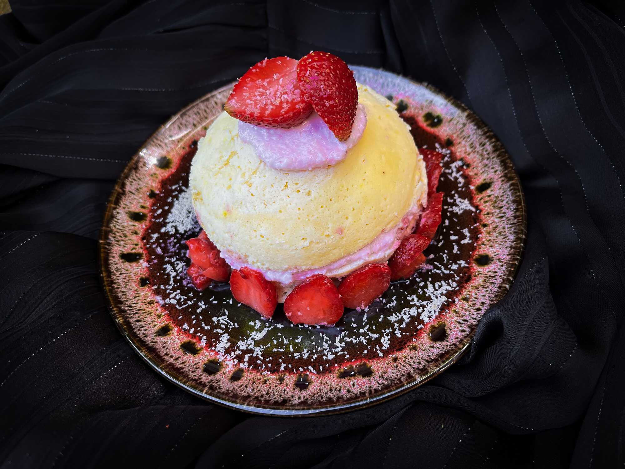 Posé sur une jolie assiette plate se trouve le bowl cake fraise et coco. Le topping est constitué avec une grosse fraise coupée en deux.