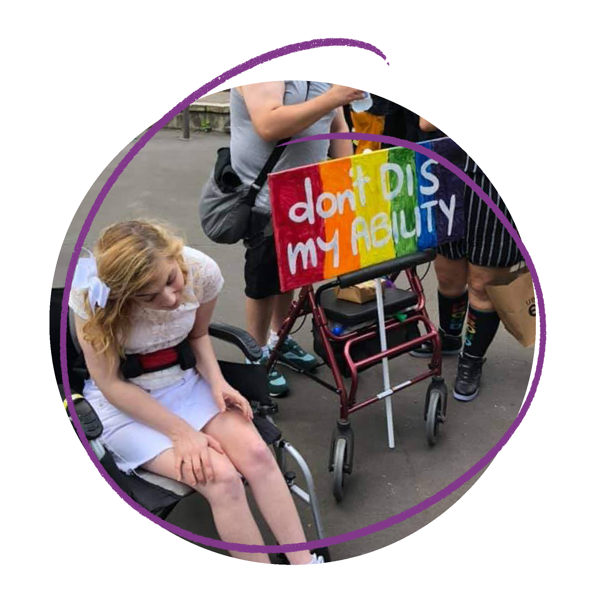 Phto of a young woman at the Mardi Gras. She is using a wheelchair and wearing a white dress. There is a person with a rainbow sign that says 'Don't DIS my ability' behind her.