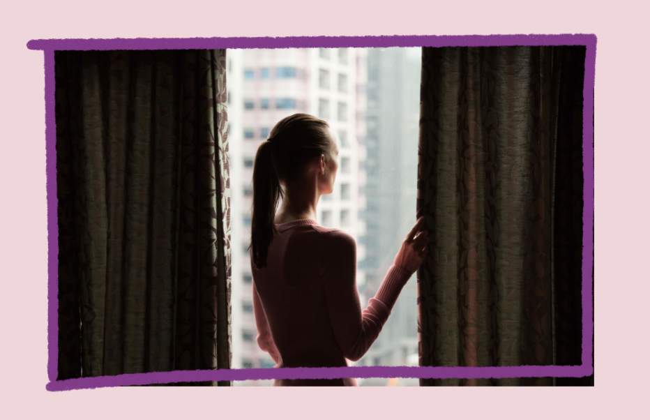 Photo of a woman with brown hair and a ponytail looking out of a window from a dark room high in a tall building.