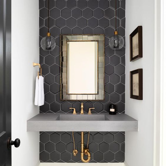 Chic black and gold powder room features clear glass pendants hung on either side of a brass tiled mirror hung from an accent wall clad in black hexagon backsplash tiles over a gray concrete sink vanity finished with a brass faucet.
