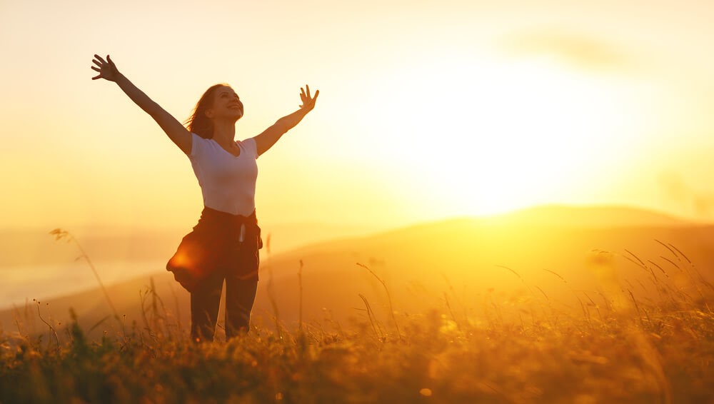 Smiling woman in a field with arms above head at sunset