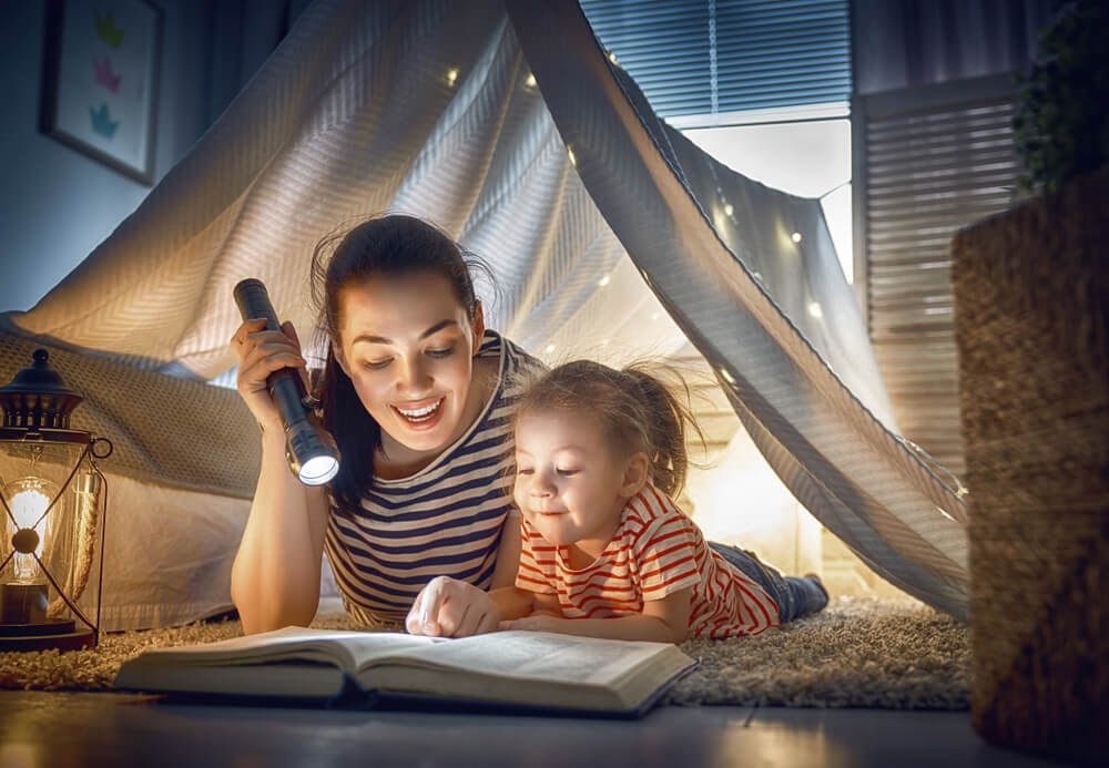 Woman and child in blanket fort reading by flashlight