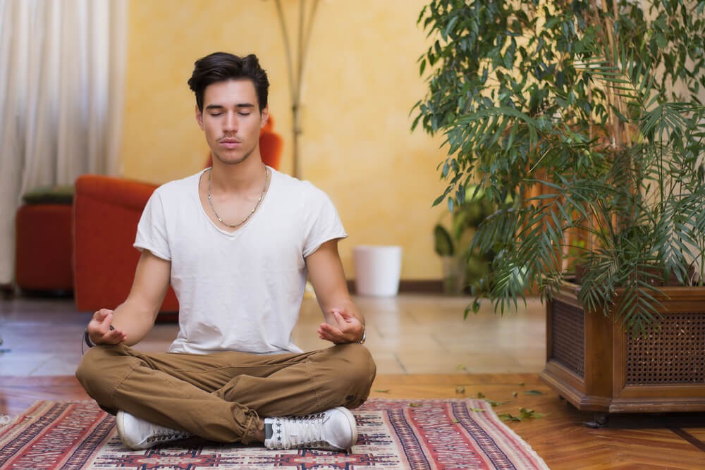 Young man sits cross legged and meditates next to potted tree
