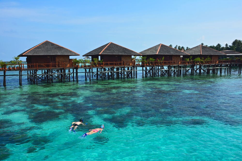 Two snorkelers in blue water below huts on stilts in the water