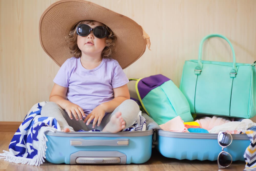 Toddler sits in packed suitcase wearings sunglasses and a big straw hat