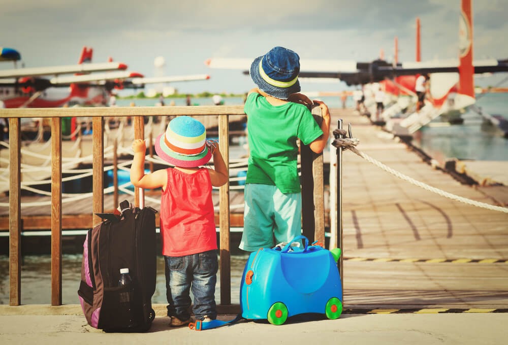 Two small toddlers look out at water planes with their suitcases. Photo is from behind.