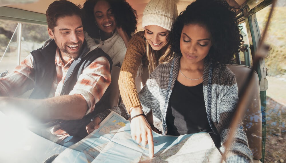 Four people are in a car looking at a map. They are happy and the person in the back seat is pointing where to go.