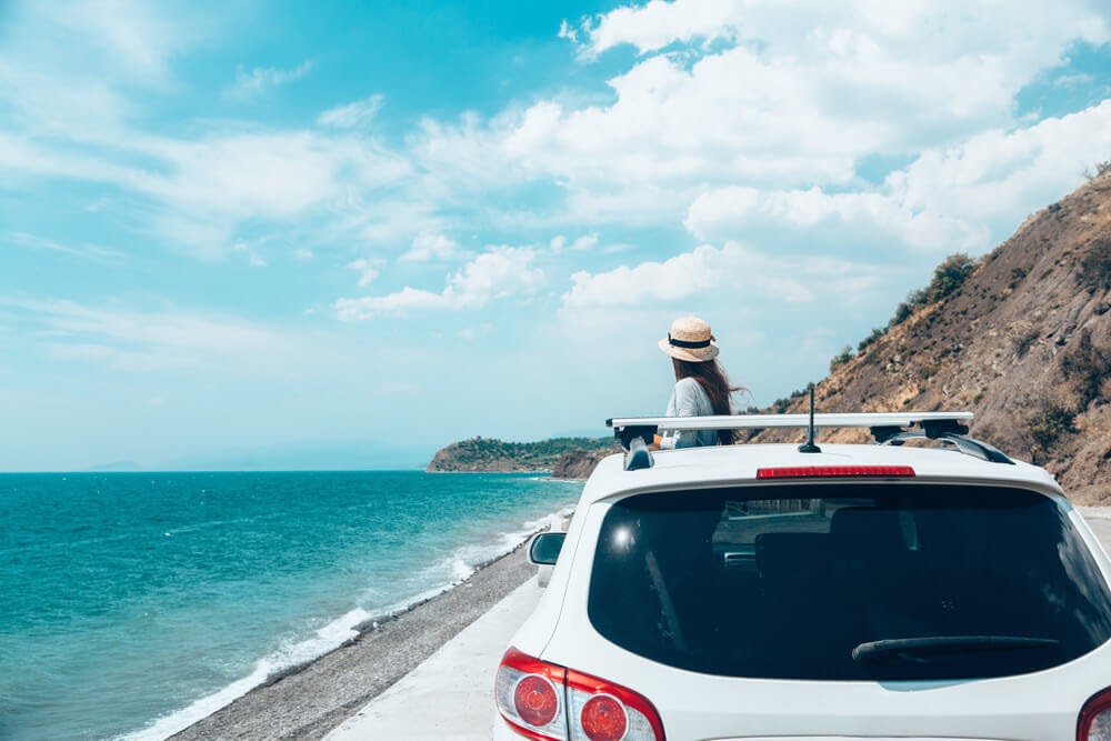 Woman standing in front of a white car along a coastal road. The sky is blue and the wind is blowing through her hair.
