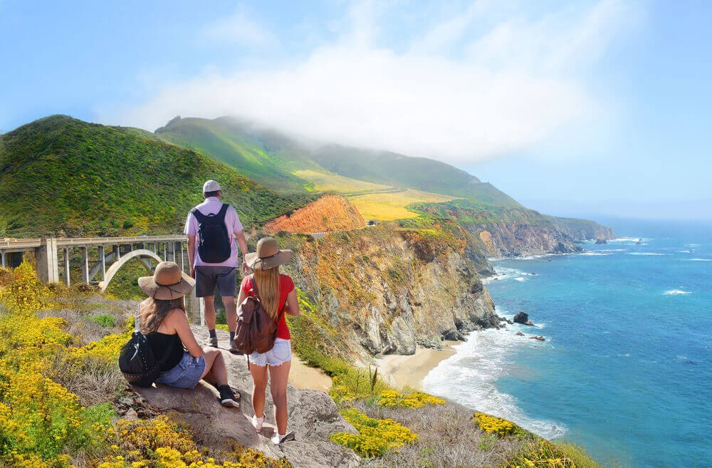 Family looks over coastline from above. They all wear backpacks and the sun is shining.